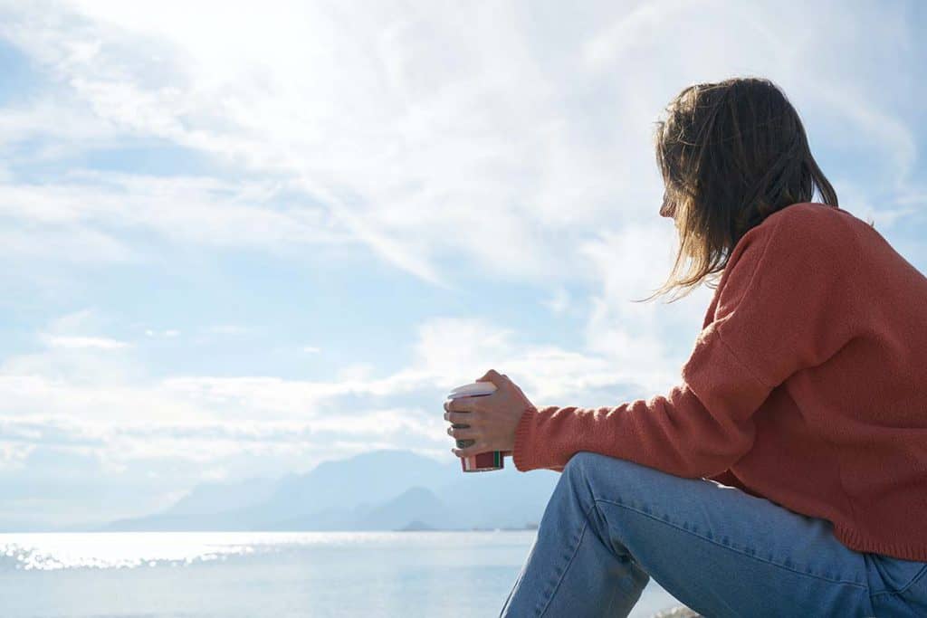 Une femme habillé d'un pull orange et de jeans regarde l'horizon. Derrière elle, il y a la mer et un ciel bleu et parsemé de nuages.