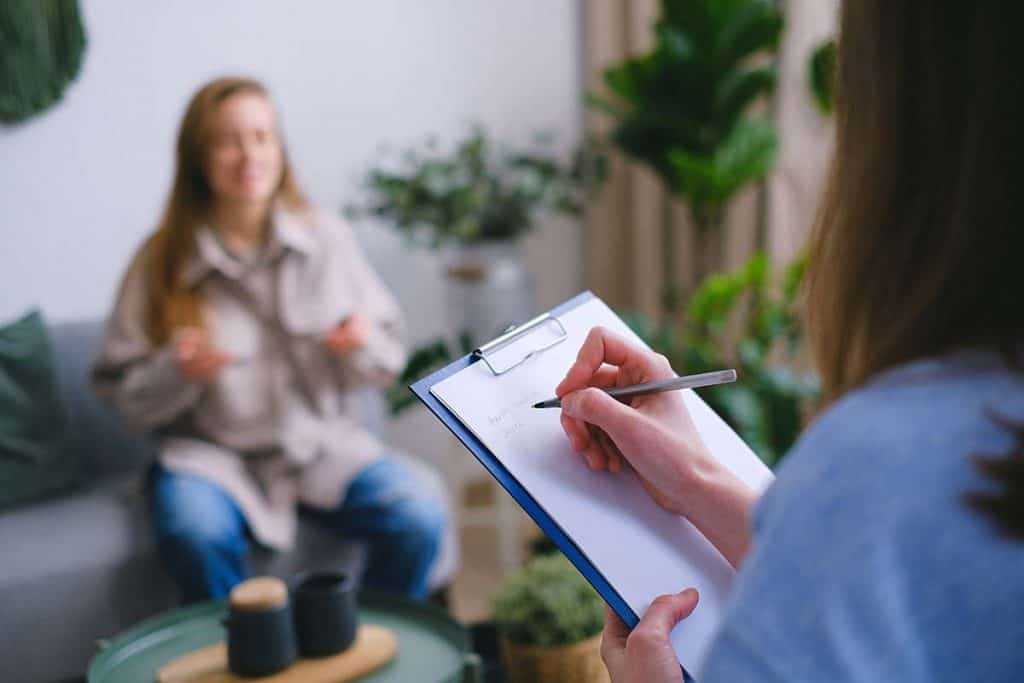 Une professionnelle habillée en bleue prend des notes. Devant elle, on voit une femme en train de parler. Elle est assise sur un sofa.