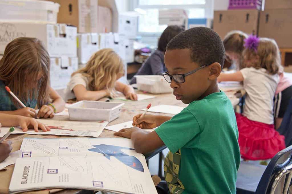 Assis à un pupitre en classe, un garçon à la peau foncée effectue un exercice dans un cahier. Il porte des lunettes et un chandail vert. On voit plusieurs autres enfants dans la pièce s'appliquer à réaliser leur exercice.