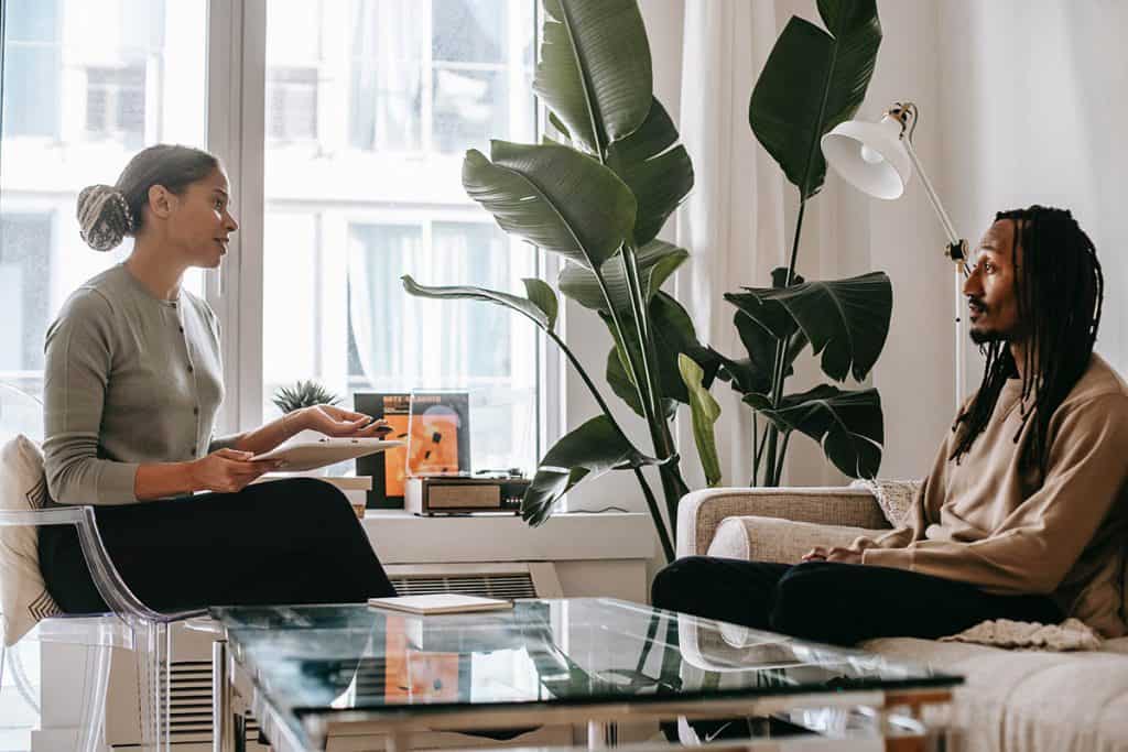 Une thérapeute parle à son client. La thérapeute est assise sur une chaise. Elle est caucasienne et porte un chandail gris et des pantalons noirs. Le client est un homme afro-américain ayant de longs cheveux. Il porte un chandail brun et des pantalons noirs. Il est assis sur un sofa. Ils sont assis sur une chaise, face à face, dans un bureau blanc. Il y a une grande plante verte et une fenêtre qui donne accès à un bloc appartement.