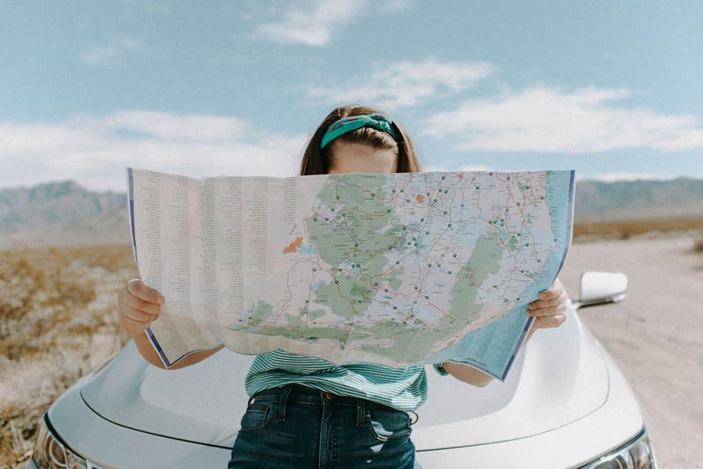 Une jeune femme regarde le plan d'une ville. Elle est assise sur le capot de sa voiture grise. On ne voit pas son visage, car il est caché par la carte. Le ciel est bleu et on y voit quelques gros nuages blancs.