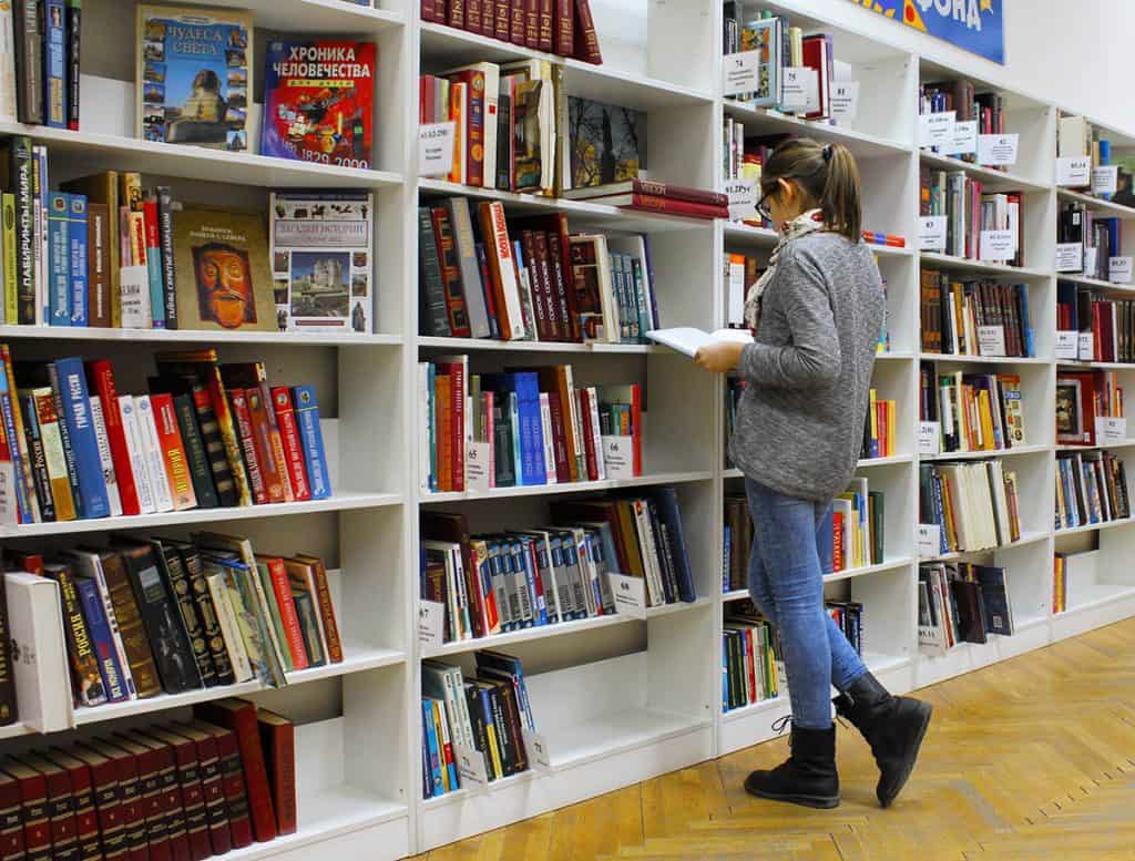 Une jeune femme feuillette un livre dans une librairie. Elle porte un chandail gris et des jeans. Elle se tient debout devant de grandes bibliothèques blanches, remplies de livres.