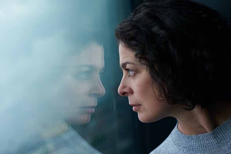 Une femme regarde à travers une fenêtre et on y voit son reflet