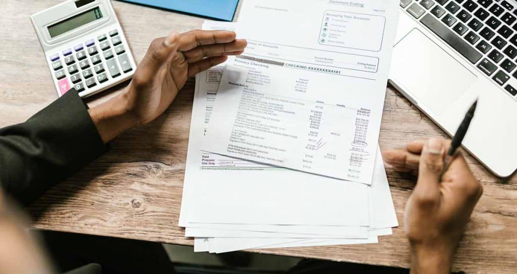 Une personne regarde des factures, un stylo à la main, entourée d'une calculatrice et d'un ordinateur portable.