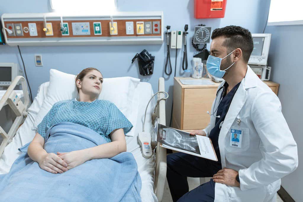 Dans une chambre d'hôpital, une patiente est couchée sur un lit et discute avec un médecin, assis juste à côté d'elle.